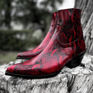 以下が含まれることがあります： 赤いヘビ柄のウエスタンブーツ。つま先が尖っていて、ブーツの後ろにジッパーが付いている。
