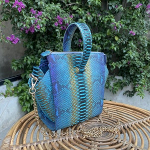 May include: A blue and gold snakeskin-patterned handbag with a top handle and a gold chain strap. The bag has a structured shape and a zip closure. The background features green foliage and purple flowers.