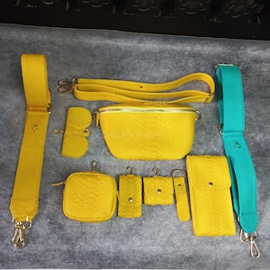 May include: A collection of yellow and teal accessories, including a fanny pack, shoulder straps, a coin purse, and a phone case. The items have a textured, snake-like pattern and gold-colored hardware. The accessories are arranged on a gray surface.