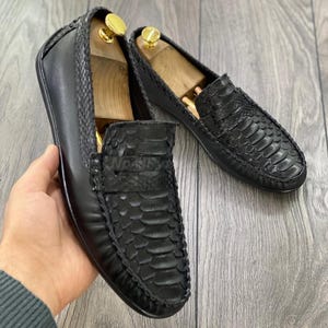Könnte beinhalten: Ein Paar schwarze Leder-Slipper mit Schlangenhautmuster. Die Slipper haben eine spitze Zehenpartie und eine Ledersohle.