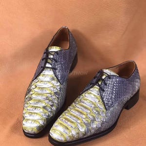 Pode incluir: Um par de sapatos de couro cinza e amarelo com padrão de pele de cobra. Os sapatos têm fecho de renda e bico pontudo.