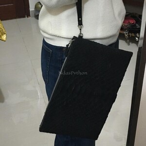 May include: Black rectangular clutch with a textured pattern, featuring a silver zipper and a black strap. The text "midasPython" is visible on the front of the clutch. The clutch is being held by a person wearing a white sweater and blue jeans.