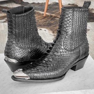 May include: A pair of black leather cowboy boots with a pointed toe and a silver metal accent on the toe. The boots are made of a textured leather that resembles snakeskin.