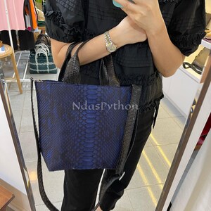 May include: A blue and black python leather tote bag with a black strap. The bag has a top handle and a long strap that can be worn over the shoulder. The bag is made of genuine python leather and has a unique, textured design.