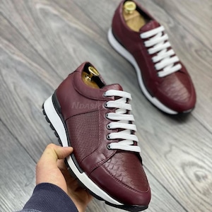 Pode incluir: Um par de tênis de couro bordô com cadarços brancos. Os tênis têm uma sola branca e uma parte superior de couro texturizada.