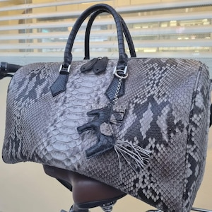 May include: A gray and black snakeskin-patterned handbag with black handles and accents. The bag features a decorative charm with a tassel and a small leather figure. The bag is sitting on a bicycle seat.