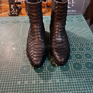 May include: A pair of black leather ankle boots with a snakeskin texture. The boots have a pointed toe, a side zip, and a small buckle detail. The boots are displayed on a green cutting mat.