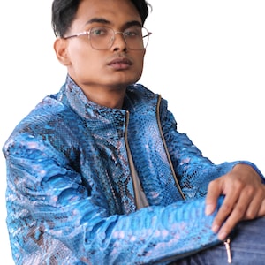 Könnte beinhalten: Eine blaue Jacke mit Schlangenmuster und Reißverschluss. Die Jacke hat eine leuchtende blaue Farbe mit dunkleren blauen und braunen Akzenten. Die Person im Bild trägt eine Brille und blaue Jeans.