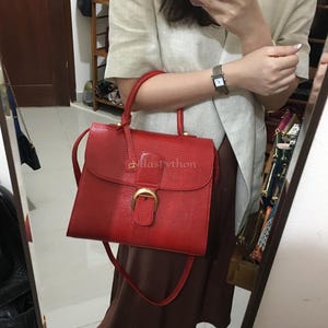 Op de afbeelding: Een rode handtas met een getextureerde afwerking, met een handvat aan de bovenkant en een schouderriem. De tas heeft een goudkleurige gespsluiting en een flap. De tekst "atlasPython" is zichtbaar op de tas.