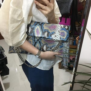 Peut inclure: Une grande pochette rectangulaire avec un motif peau de serpent dans les tons de bleu, rose et jaune. La pochette a une chaîne argentée et un rabat. Le sac est tenu par une personne.