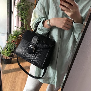 May include: A black handbag with a snakeskin pattern, a top handle, and a silver buckle closure. The bag has a long, adjustable strap. The text "NdasPython" is visible on the front of the bag.