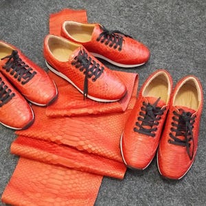 Könnte beinhalten: Vier Paar rote Schlangenlederschuhe mit schwarzen Schnürsenkeln. Die Schuhe sind auf einem roten Schlangenlederstoff angeordnet.