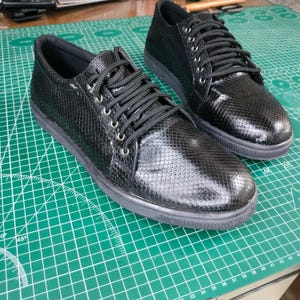 Pode incluir: Um par de tênis de couro preto com um padrão texturizado de pele de cobra. Os cadarços são pretos e os tênis têm uma sola de borracha branca.