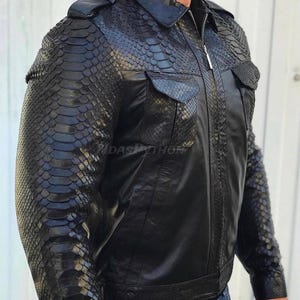 May include: A black leather jacket with a snakeskin pattern on the sleeves and pockets. The jacket has a zip fastening and a collar.