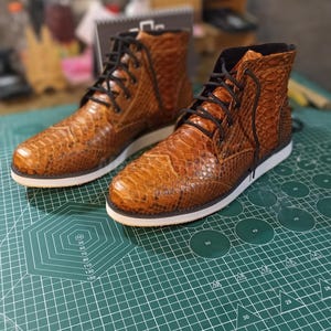 May include: Two brown leather boots with a snakeskin pattern and white soles. The boots are laced up and have a high top.