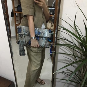 Puede incluir: Un bolso de mano con estampado de piel de serpiente con detalles azules y una cadena plateada. El bolso tiene una borla y se sostiene frente a un espejo. Un accesorio de moda.