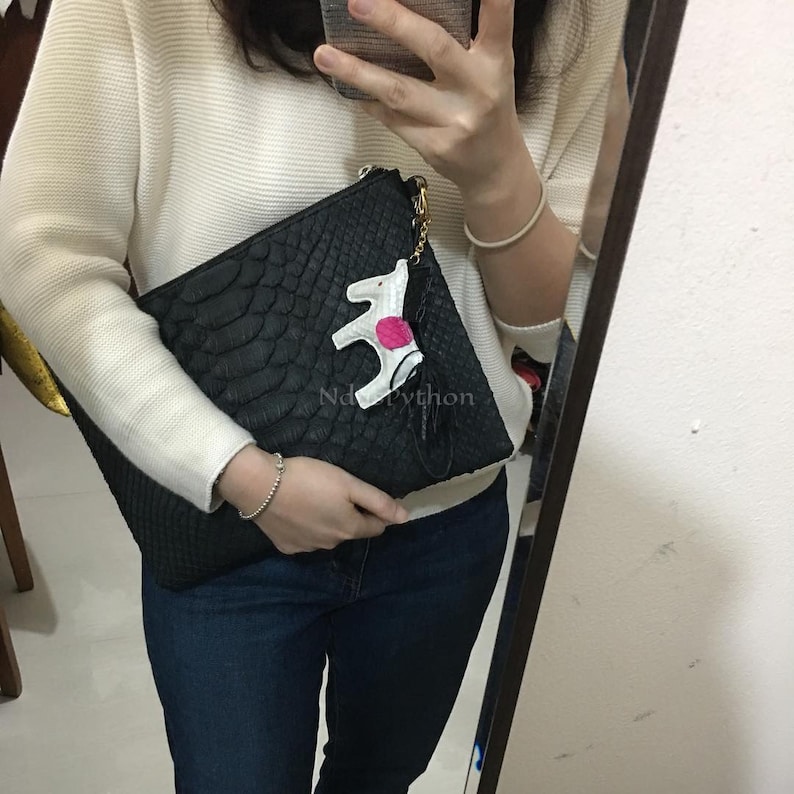May include: Black clutch with a snakeskin texture, featuring a white and pink decorative charm. The clutch has a gold zipper and a small tag that reads "Nd Python". The bag is held in front of a mirror.