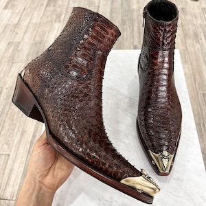 Könnte beinhalten: Ein Paar braune Cowboy-Stiefel aus Leder mit spitzer Zehenkappe und einem goldenen Metallakzent an der Spitze. Die Stiefel sind aus strukturiertem Leder, das an Schlangenhaut erinnert.