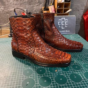 Könnte beinhalten: Braune Leder-Ankle Boots mit Reptilienhaut-Textur. Die Stiefel haben einen Reißverschluss und eine eckige Zehenpartie. Sie werden auf einer grünen Schneidematte präsentiert. Der Text "ALAPATI BALI" ist im Hintergrund sichtbar.