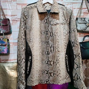 Pode incluir: Uma jaqueta de couro de cobra marrom e bege com fecho de zíper. A jaqueta tem um painel preto de cada lado e um bolso do lado esquerdo.