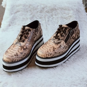 May include: A pair of platform shoes with a snakeskin pattern in shades of brown and tan. The shoes have thick white and black striped soles, brown laces, and a zipper detail on the side.