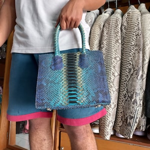 Puede incluir: Un bolso de mano de piel de serpiente azul, verde y negro con asa superior. El bolso tiene un patrón texturizado y escamoso.