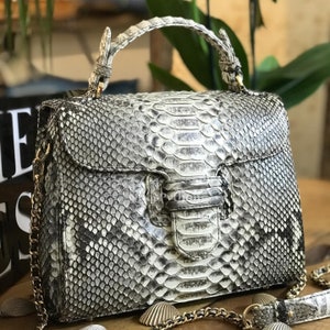 Puede incluir: Un bolso de mano de piel de serpiente blanco y gris con una correa de cadena dorada y un asa superior. El bolso tiene un cierre de solapa con una hebilla dorada.