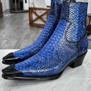 Puede incluir: Un par de botines azules con textura de piel de serpiente, con punteras y tacones negros. Los botines tienen un diseño de punta afilada y una cremallera lateral. Están hechos de un material texturizado con un ligero brillo.