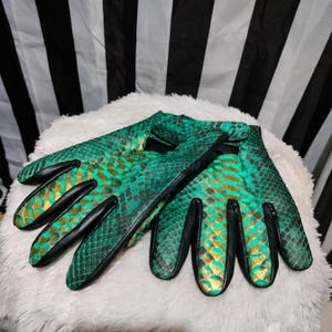 Peut inclure: Une paire de gants en peau de serpent vert et or avec des finitions en cuir noir. Les gants sont pliés en deux et les doigts sont écartés.