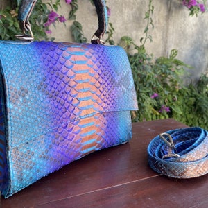 Peut inclure: Sac à main en peau de serpent bleu et violet avec une bandoulière assortie. Le sac a une poignée supérieure et une fermeture à rabat. Le motif peau de serpent présente des nuances de bleu, de violet et de bronze.