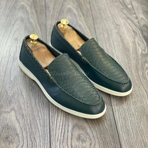 May include: A pair of dark green leather loafers with a snakeskin pattern. The loafers have white soles and are made of a soft, comfortable material.