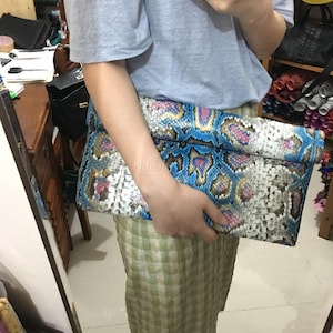 May include: A rectangular clutch with a snakeskin pattern in shades of blue, pink, and white. The clutch is held by a person. The pattern resembles a snake's scales. The bag is a fashion accessory.