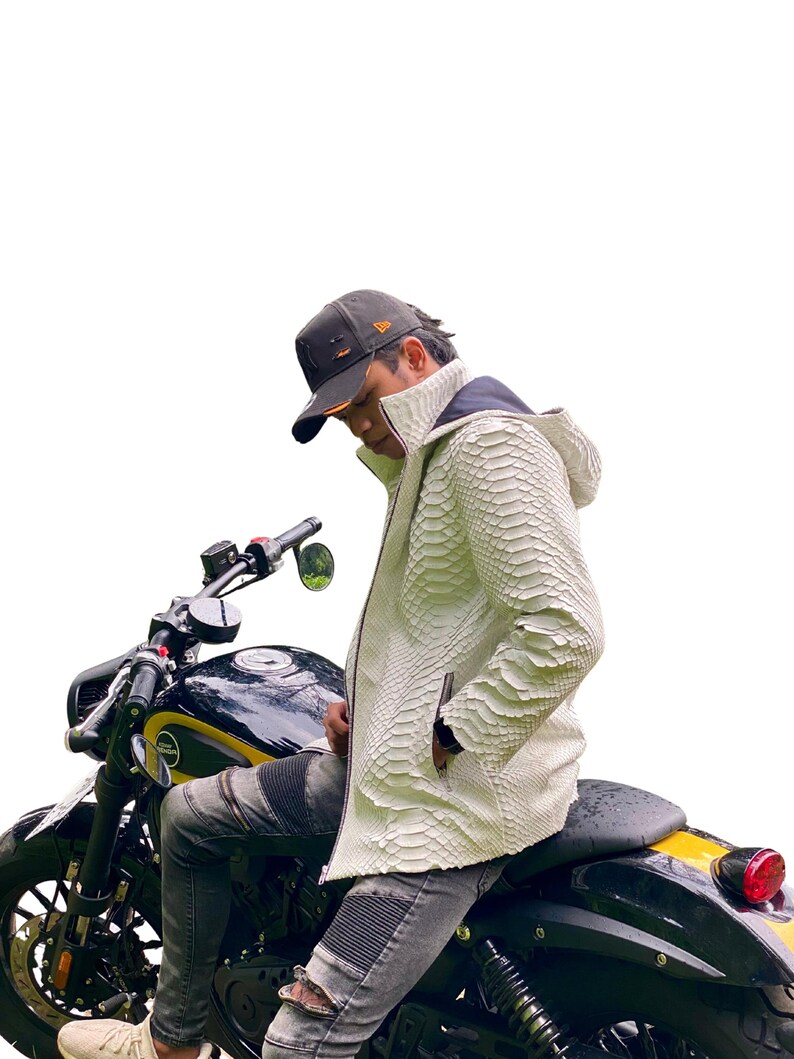 May include: A white, textured leather jacket with a hood and zip closure. The jacket is worn over a black and yellow motorcycle. The person is wearing a black baseball cap and grey jeans.