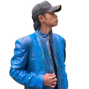 Pode incluir: Casaco azul com padrão de pele de cobra e fecho de correr. O casaco tem gola alta e mangas compridas. A pessoa na imagem está a usar um boné de beisebol cinzento com viseira preta.