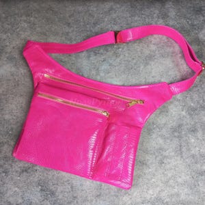 Op de afbeelding: Een felroze heuptasje met een textuurpatroon dat op reptielen lijkt. De tas heeft twee compartimenten met ritssluiting en een kleinere zak, allemaal met goudkleurige ritsen. De verstelbare riem en gesp zijn ook roze.