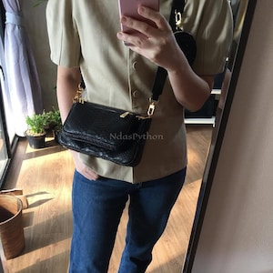 Puede incluir: Bolso bandolera negro con un patrón texturizado, herrajes dorados y una larga correa negra. El bolso se lleva al hombro. El texto "NdasPython" es visible en el bolso.