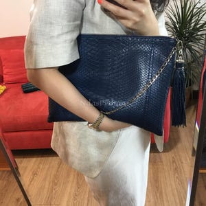 Puede incluir: Un bolso de mano azul marino con un estampado de piel de serpiente, una cadena dorada y una borla a juego. La bolsa rectangular es sostenida por una persona. El fondo incluye un sofá rojo, una planta y un espejo.