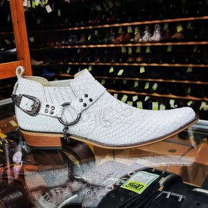 Op de afbeelding: Witte leren cowboylaarzen met een spitse neus en een decoratieve gesp en ketting. De laarzen hebben een getextureerd, slangachtig patroon.