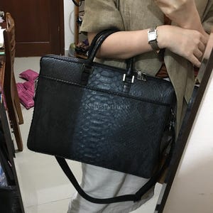 May include: Black briefcase with a snakeskin texture. The bag features a top handle, a shoulder strap, and a front pocket. The text "NedsPython" is visible on the front. The bag is held by a person.