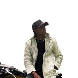 May include: A white, textured leather jacket with a snake-skin pattern. The jacket has a zip closure and a high collar. The person in the image is wearing a black baseball cap and a black shirt. The person is sitting on a motorcycle.