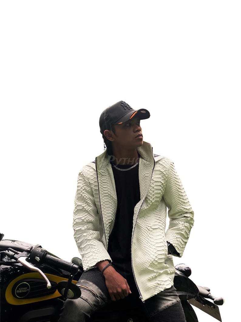 May include: A white, textured leather jacket with a snake-skin pattern. The jacket has a zip closure and a high collar. The person in the image is wearing a black baseball cap and a black shirt. The person is sitting on a motorcycle.
