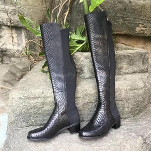 Pode incluir: Um par de botas pretas de cano alto com padrão de pele de cobra. As botas têm um salto bloco e uma ponta pontiaguda.