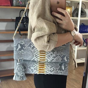 Peut inclure: Pochette en peau de serpent grise et blanche avec une bandoulière chaîne dorée et un pompon assorti. La pochette a un rabat et une bande centrale d'écailles dorées. D'autres sacs à main sont visibles sur des étagères.