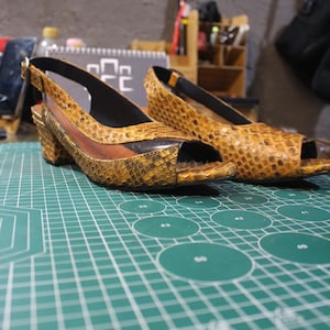 Puede incluir: Un par de sandalias de tacón de piel de serpiente marrón con puntera abierta y correa de slingback. Las sandalias están sobre una estera de corte verde con círculos blancos.