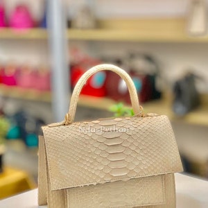 May include: A beige python-skin handbag with a structured design. The bag features a top handle, a flap closure, and a front pocket. The text "NdasPython" is visible on the front. The bag is displayed on a white surface.