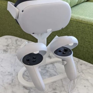 May include: A white VR headset stand with two white controllers. The stand has a white base and a white tree-like structure that holds the headset and controllers.