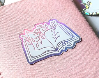 Può includere: Un adesivo olografico di un libro aperto con foglie e viti su uno sfondo di tessuto rosa.
