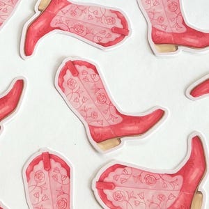 May include: A collection of red cowboy boot stickers with a floral design. The boots have a light pink floral pattern on the shaft and foot. The stickers are arranged on a white background.