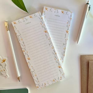 May include: Two white notepads with lined paper and a floral design featuring yellow flowers and green stems. The notepads have the word "notes" printed at the top.