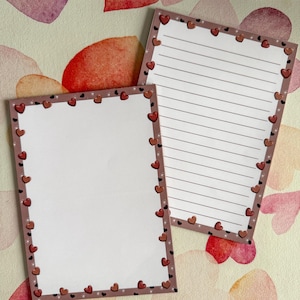 Pode incluir: Dois blocos de notas retangulares com uma borda de pequenos corações e estrelas vermelhas. Um bloco de notas tem páginas pautadas, o outro está em branco. Os blocos de notas estão em um fundo com corações em aquarela em tons de rosa e vermelho.